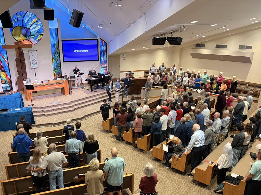Grace Lutheran Church congregation standing in worship beneath the sanctuary's stained-glass window and cross.