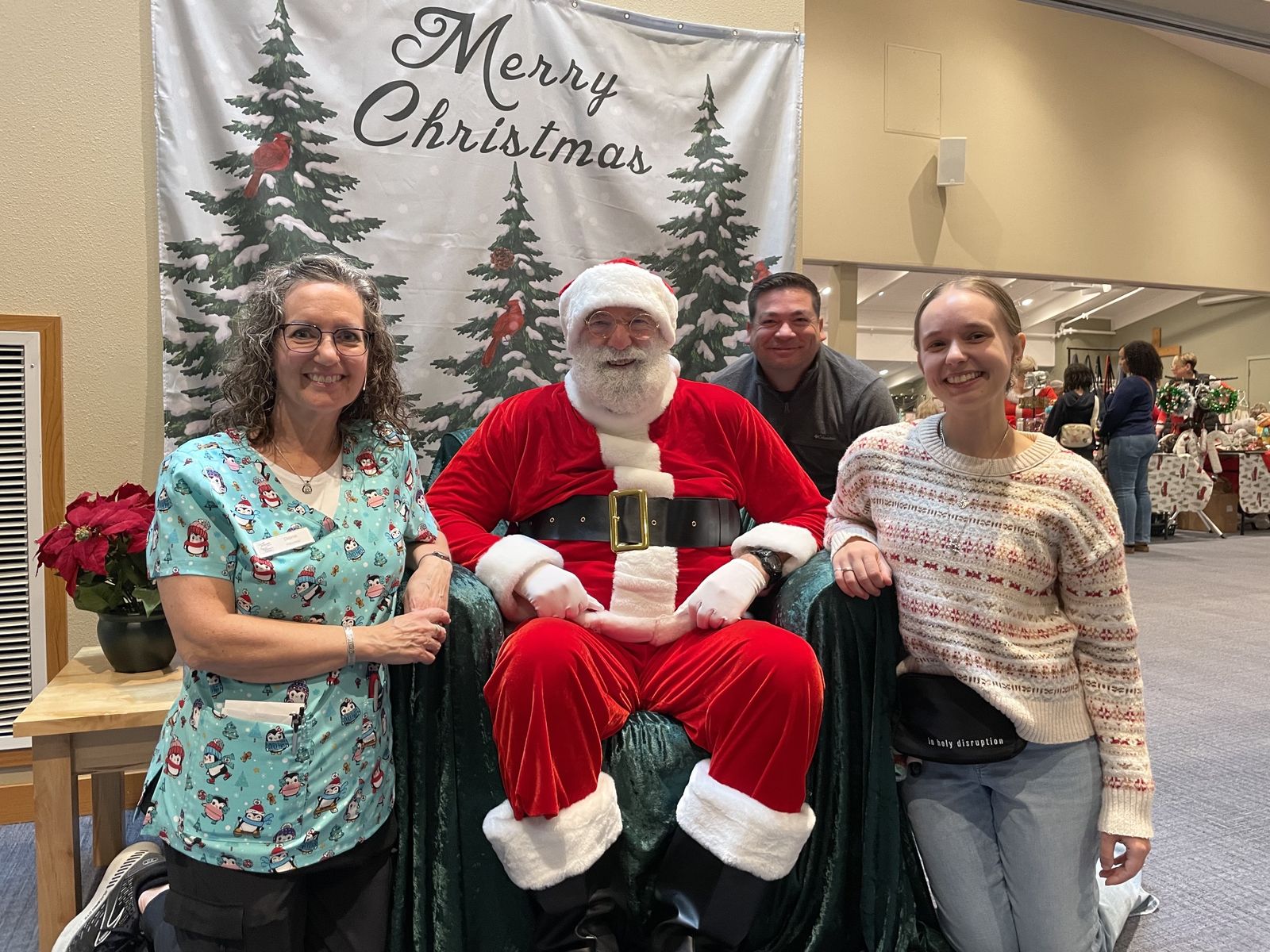 Santa seated in front of a Merry Christmas backdrop with three smiling volunteers